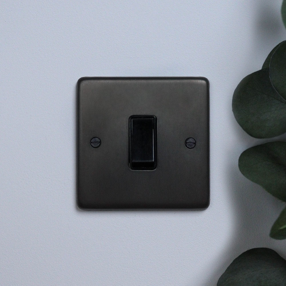 A dark bronze 1 gang 2 way electrical switch against a light background, with a plant to the right of the switch.