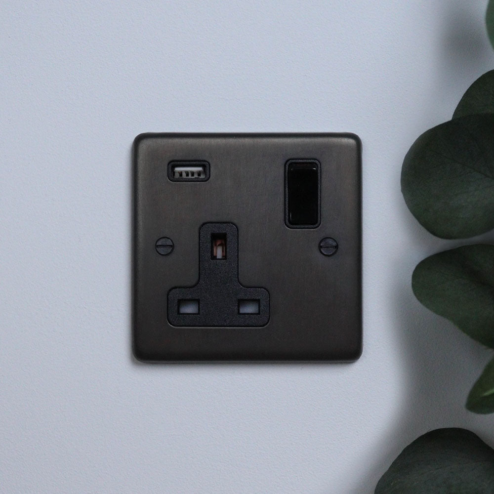 A dark bronze single socket with a USB port, mounted on a white wall next to a green potted plant.